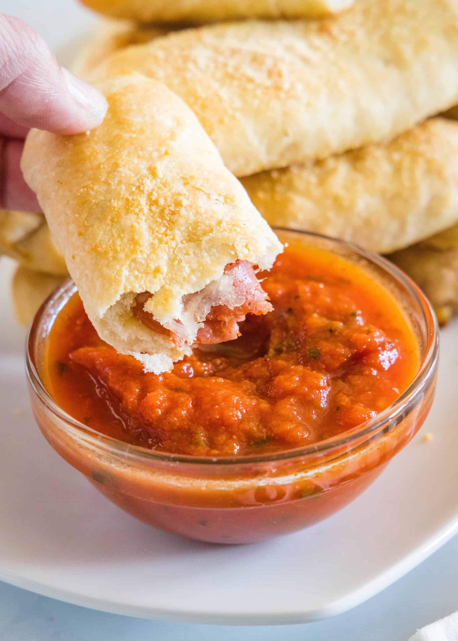 A hand holding one half of a pepperoni roll above a small bowl of marinara sauce with more rolls in the background.
