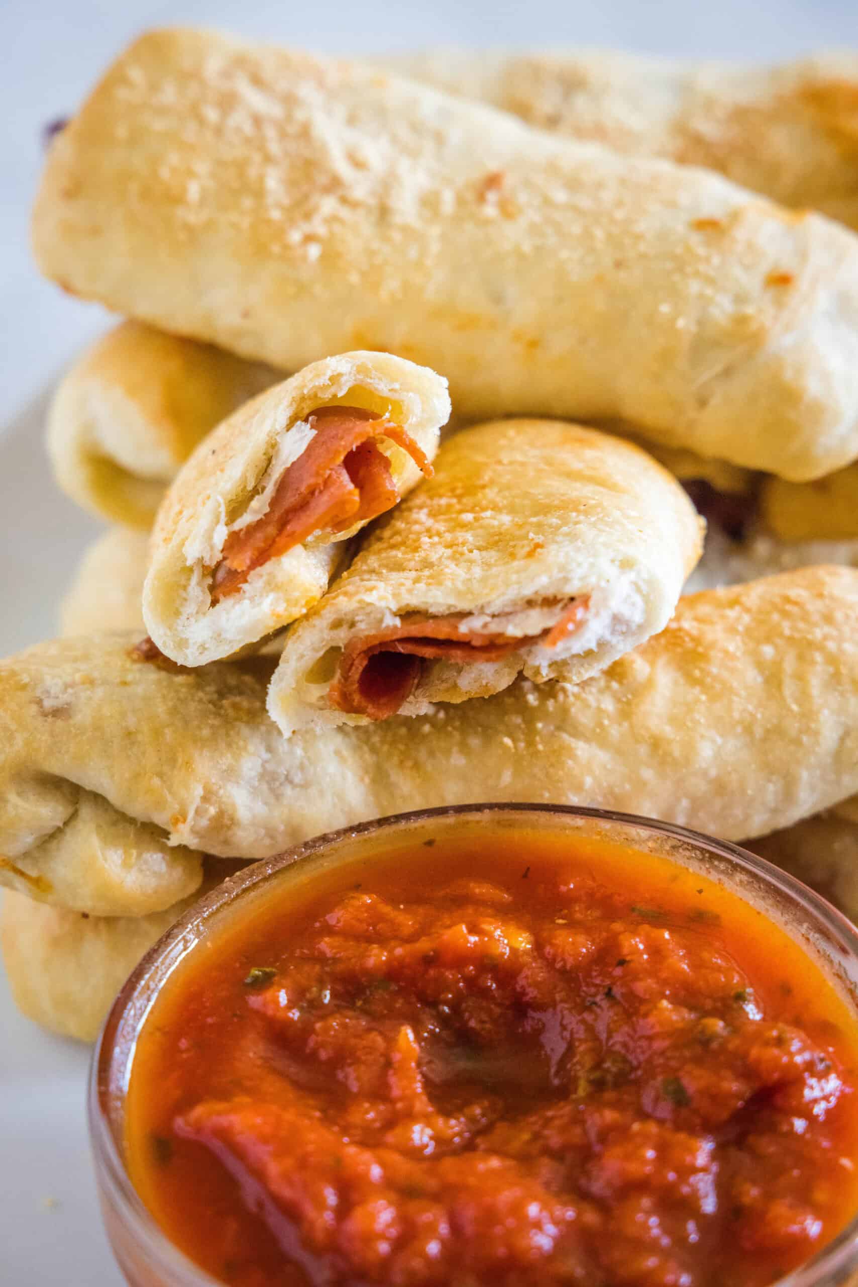 These pepperoni rolls are an easy snack made from soft, fluffy pizza dough filled with melty mozzarella cheese and juicy pepperoni slices. Pepperoni rolls stacked behind a bowl of marinara sauce, with one roll broken in half.