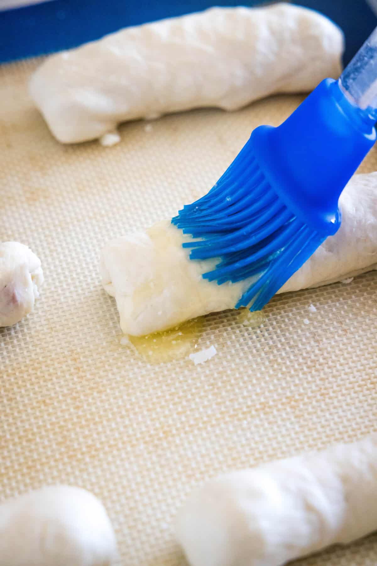 A blue rubber basting brush coating the top of an unbaked pepperoni roll with melted butter.