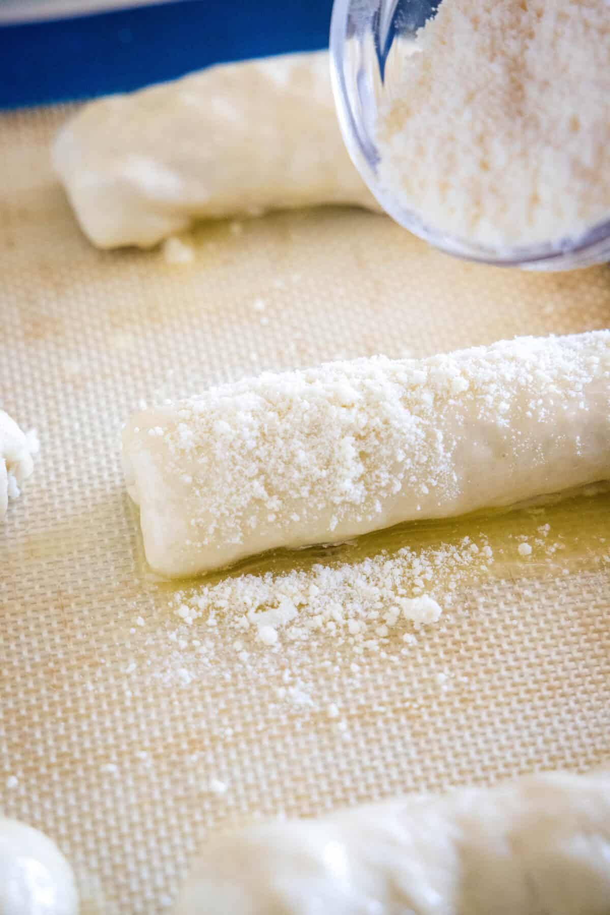 Grated parmesan being sprinkled from a bowl over top prepared pepperoni rolls on a lined baking sheet.