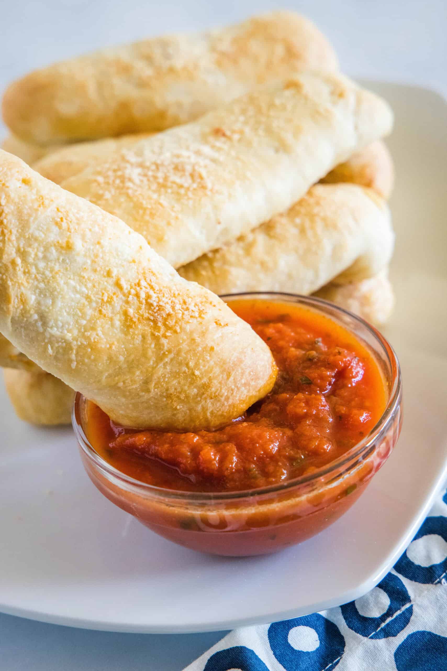A pepperoni roll being dipped into a small bowl of marinara sauce with more rolls in the background.