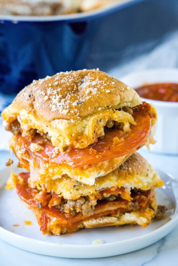 Two pizza sliders stacked on a white plate with a bowl of marinara sauce in the background.