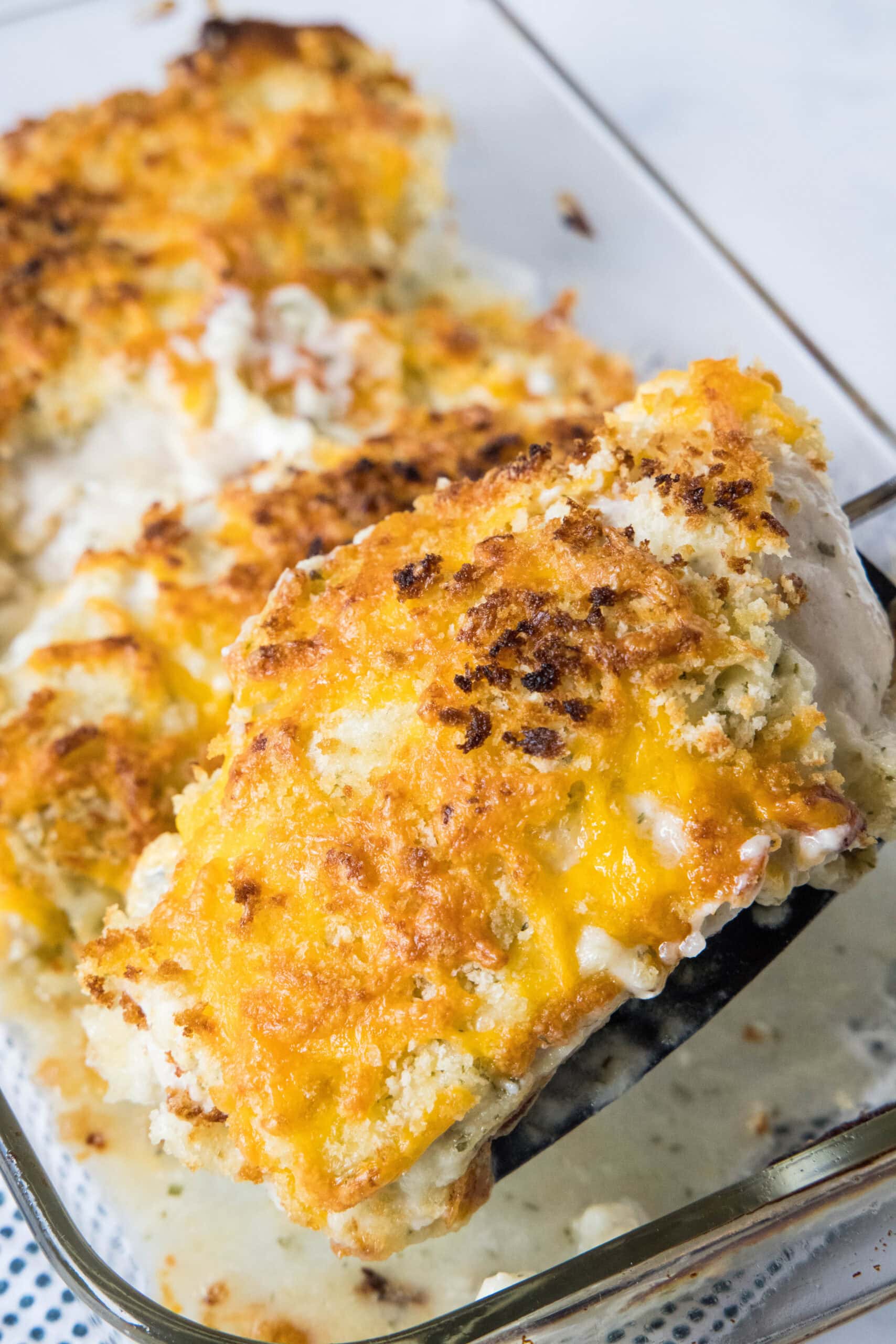 A spatula lifting a cheesy ranch chicken breast from a glass baking dish.