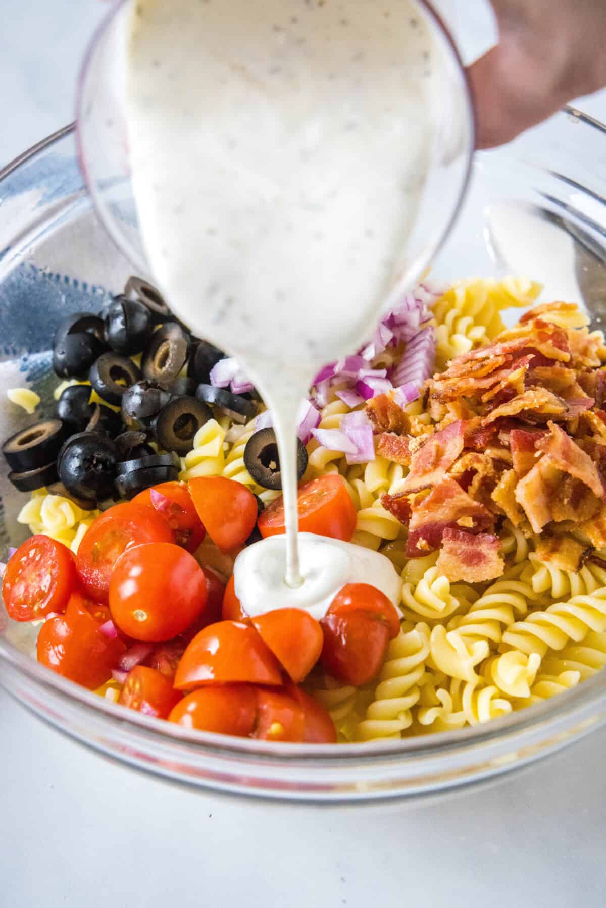 Ranch dressing being poured over pasta salad ingredients in a glass bowl.