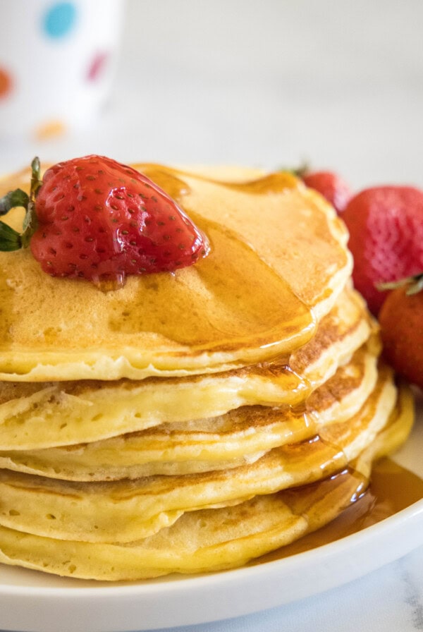 A stack of ricotta pancakes topped with a strawberry and maple syrup on a plate, next to more fresh strawberries.