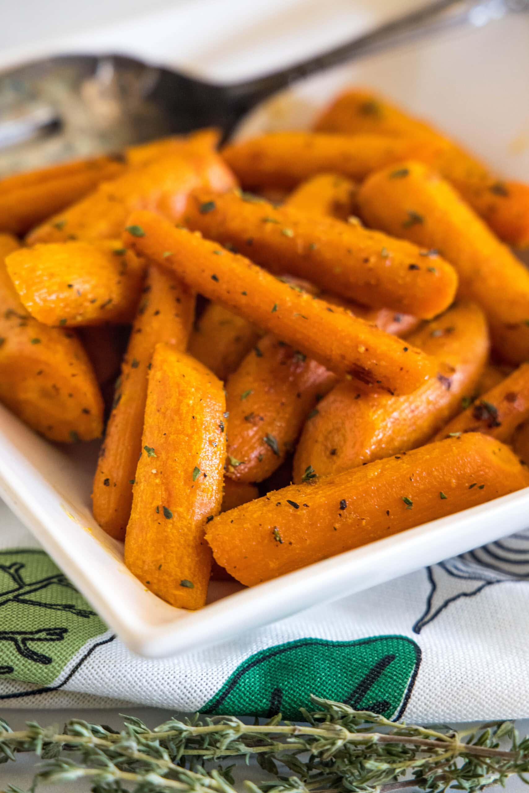 Seasoned roasted carrots in a square white bowl with a serving spoon.