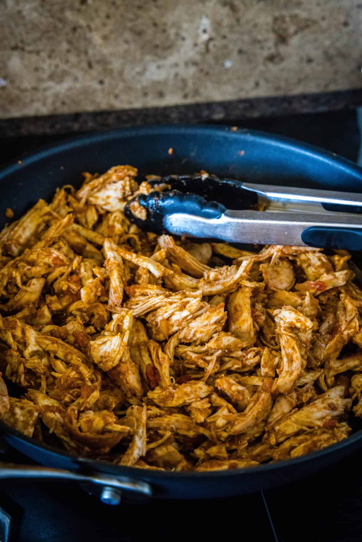 Shredded salsa rotisserie chicken in a skillet with a set of tongs.