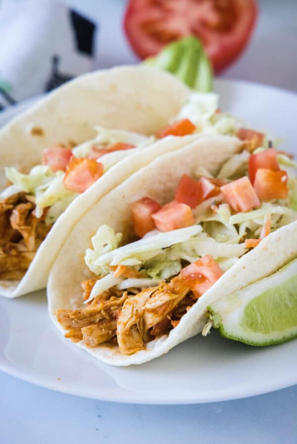 Two rotisserie chicken tacos topped with Mexican coleslaw and diced tomatoes on a white plate, next to a lime wedge.