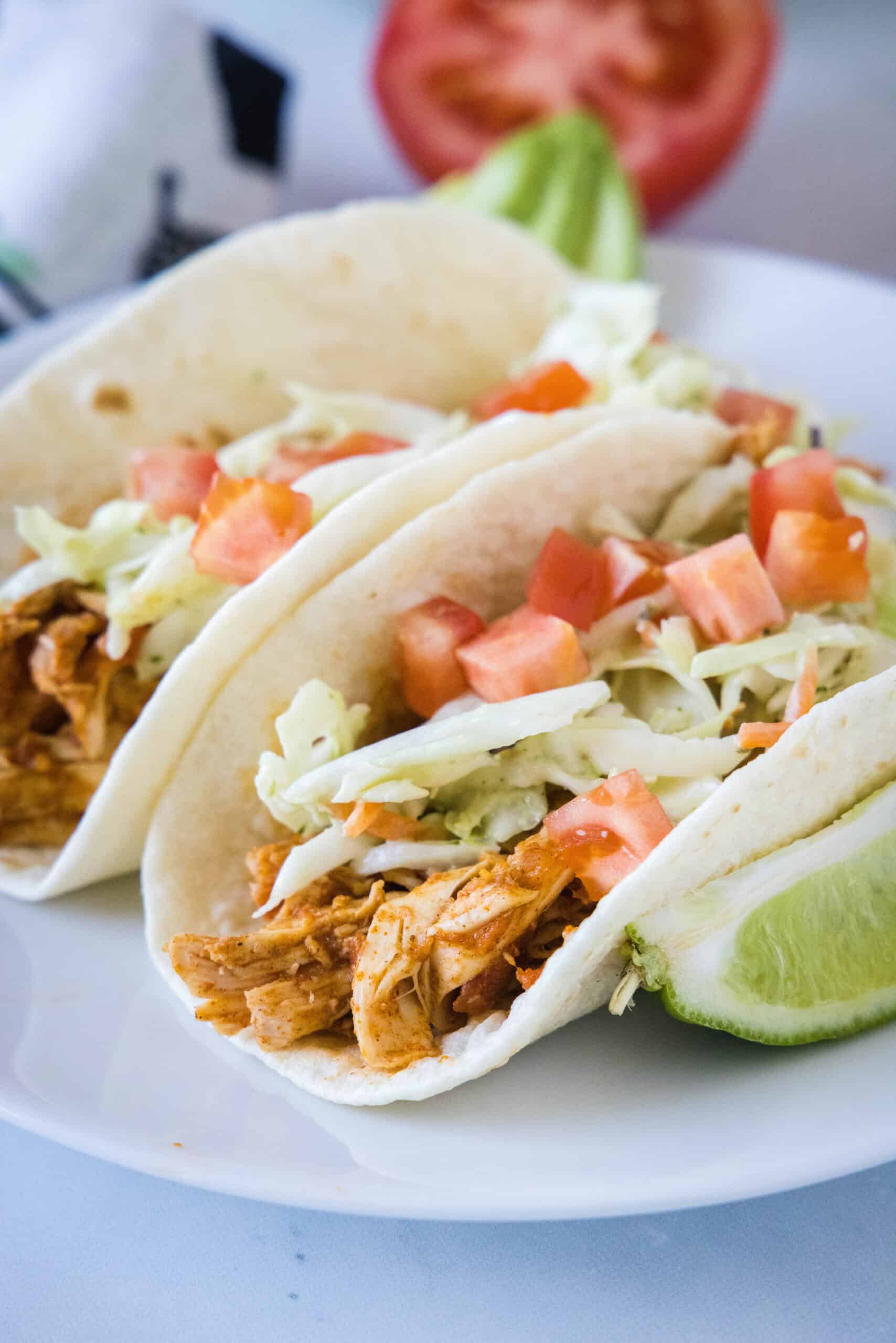 Two rotisserie chicken tacos topped with Mexican coleslaw and diced tomatoes on a white plate, next to a lime wedge.