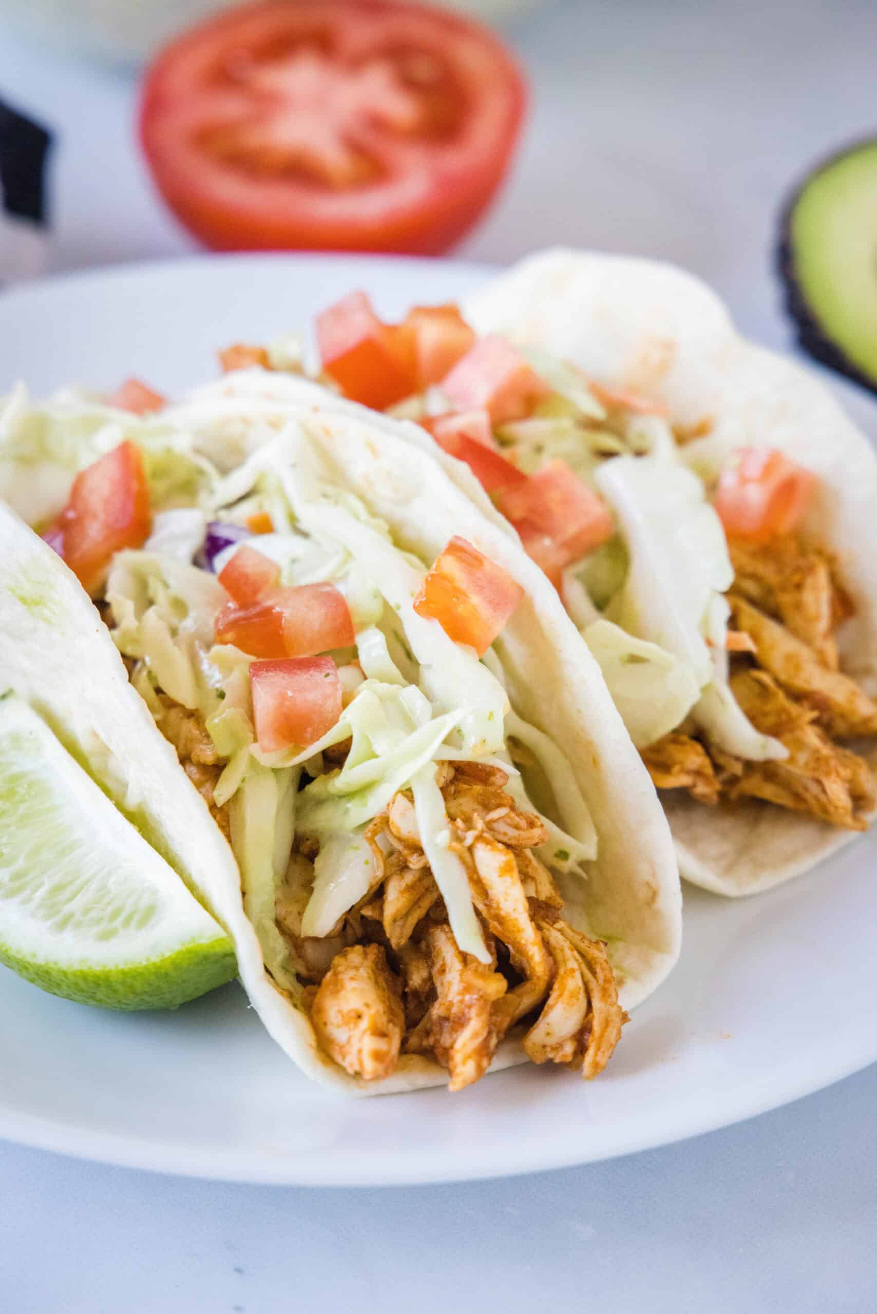 Two rotisserie chicken tacos topped with Mexican coleslaw and diced tomatoes on a white plate, next to a lime wedge.