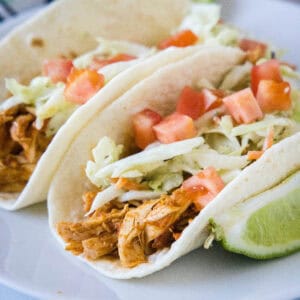 Two rotisserie chicken tacos topped with Mexican coleslaw and diced tomatoes on a white plate, next to a lime wedge.