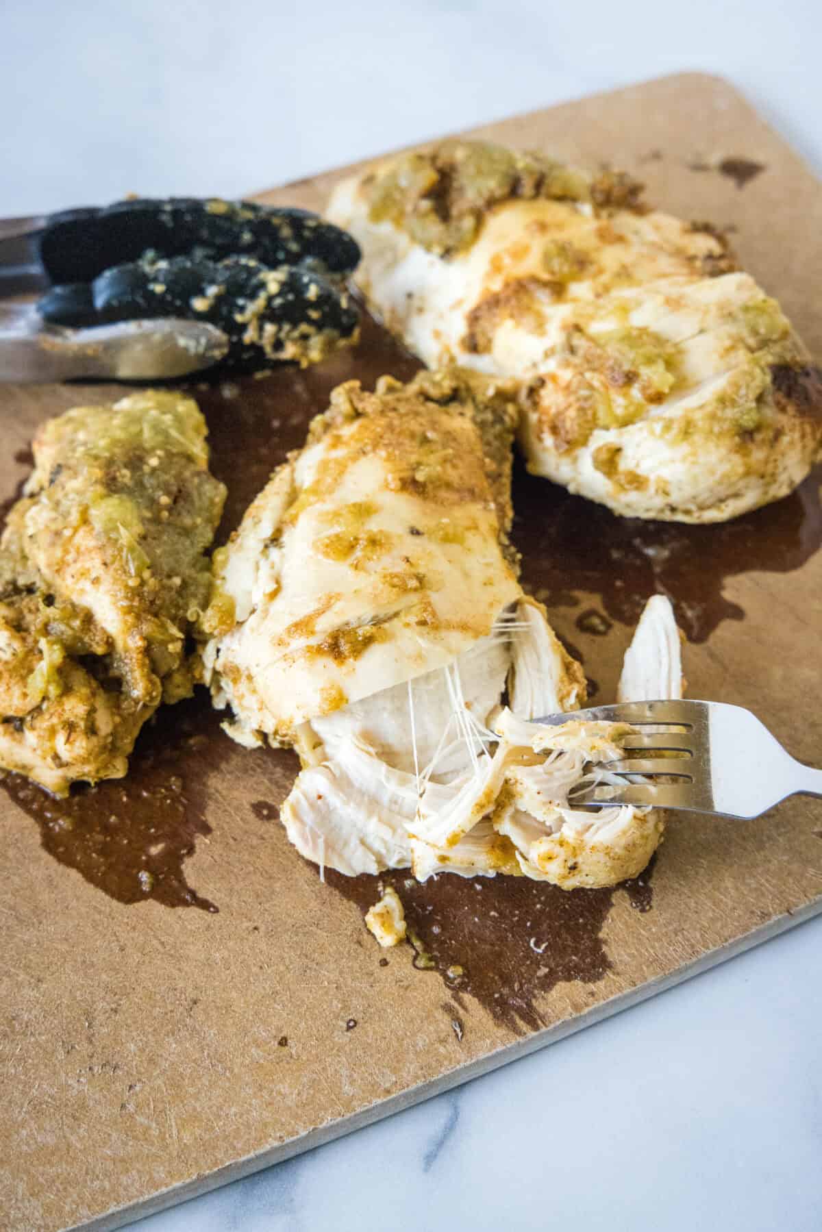 A fork shredding a salsa verde chicken breast on a cutting board.