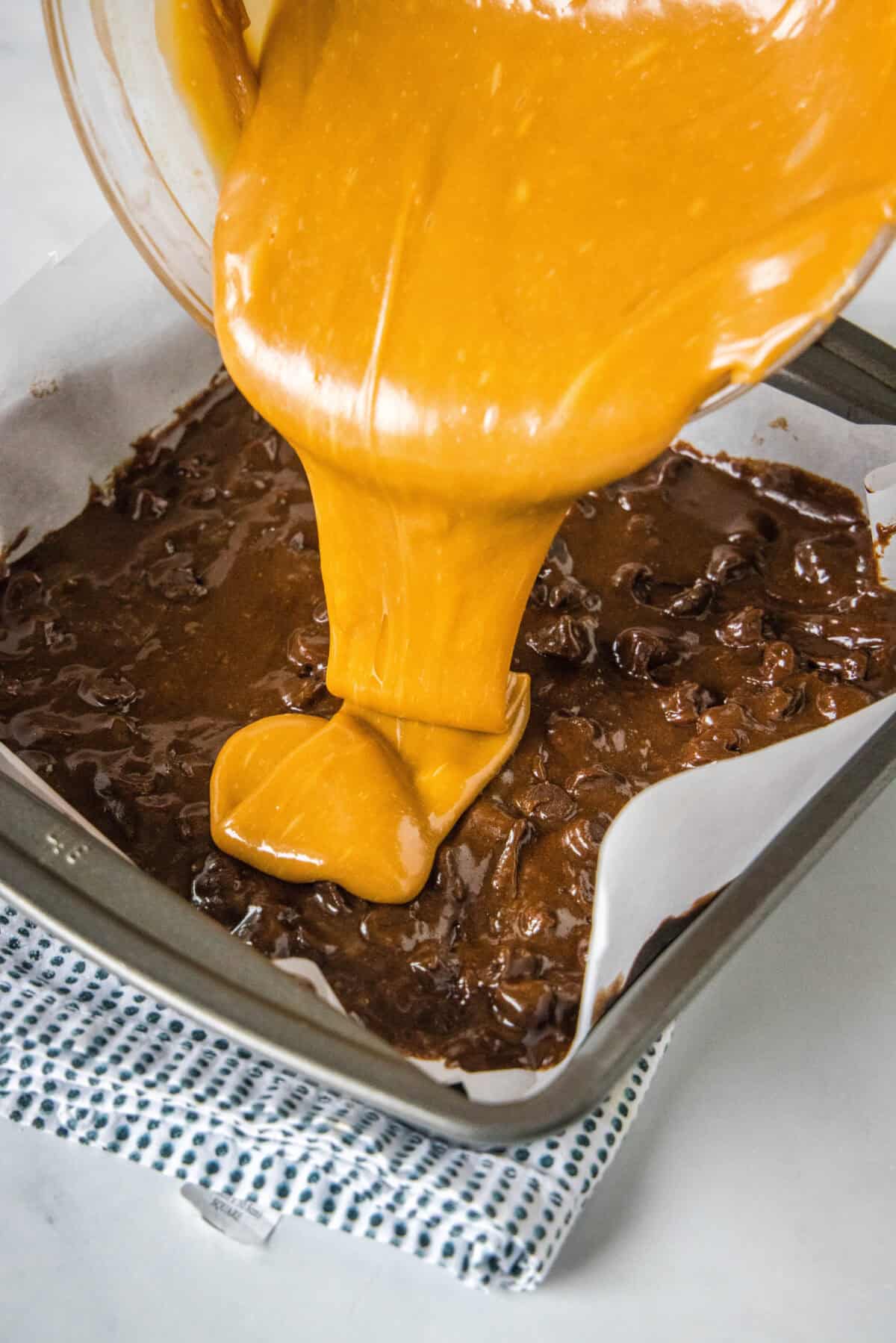 Salted caramel being poured over brownie batter in a lined square pan.