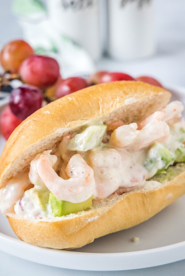A shrimp salad sandwich served on a white plate with red grapes in the background.