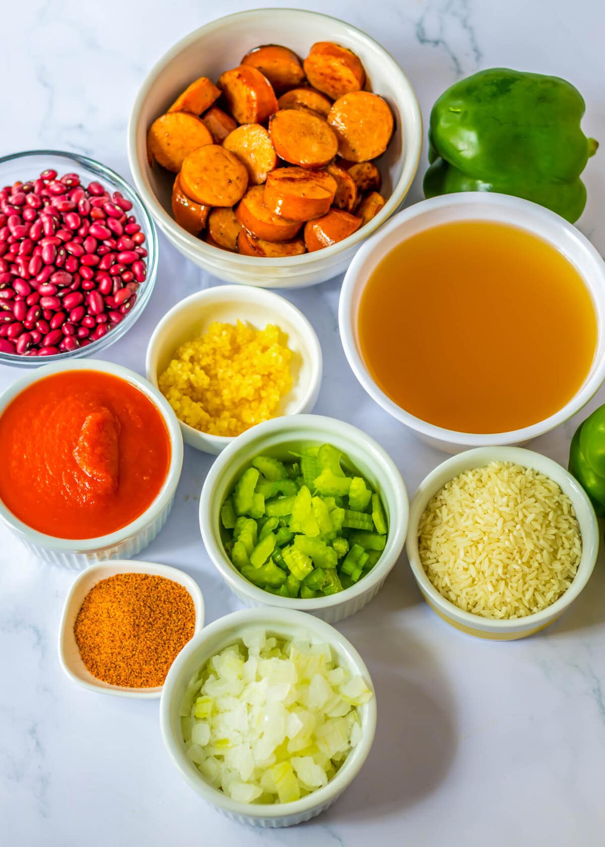 Overhead view of the ingredients needed for red beans and rice: a bowl of sliced sausage, a bowl of red beans, a bowl of tomato sauce, a bowl of chicken broth, a bowl of diced celery, a bowl of diced onion, a bowl of minced garlic, a bowl of rice, a bowl of cajun seasoning, and a whole green bell pepper.