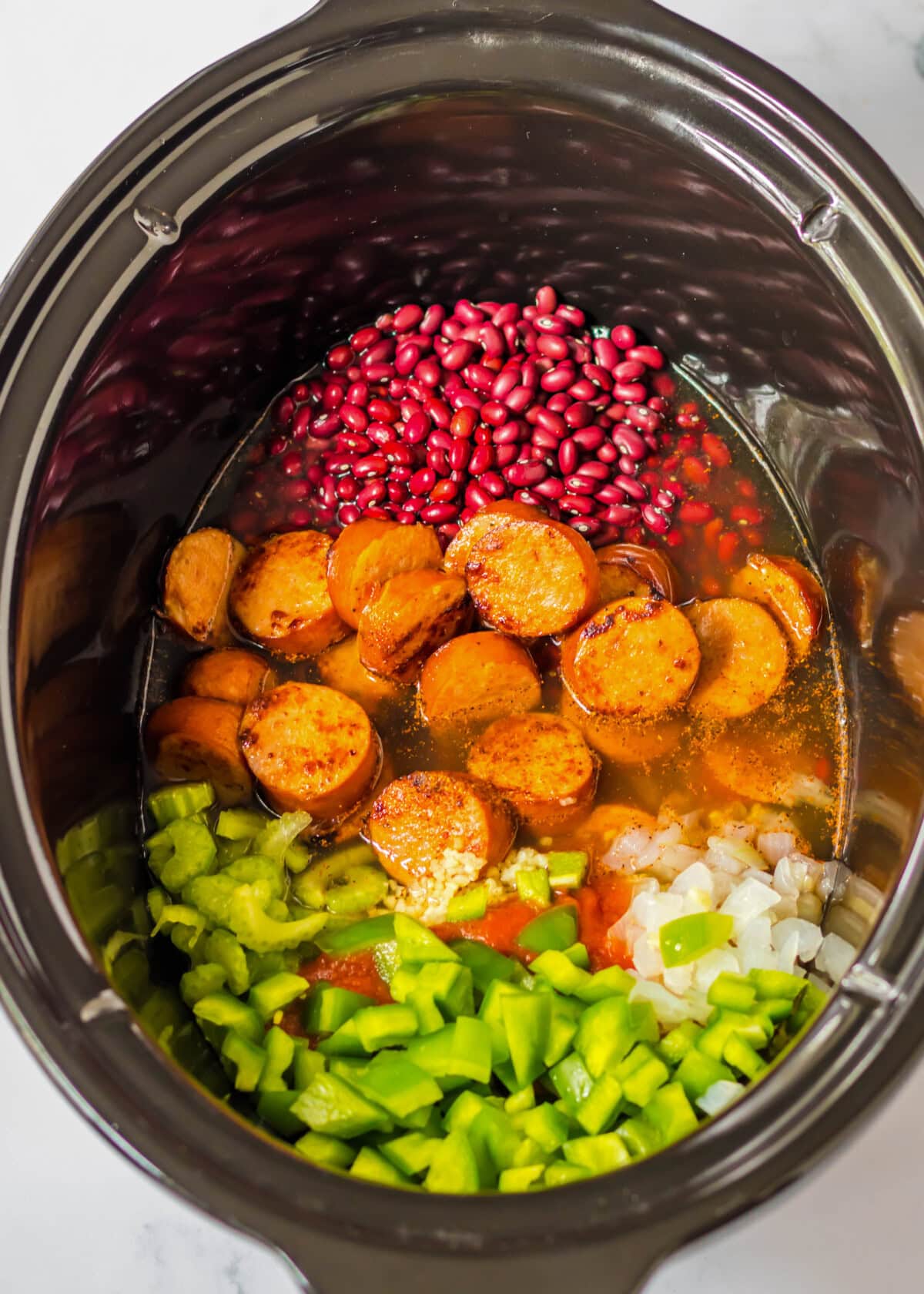 A crock pot filled with dried red beans, sliced sausage, celery, onions, green bell pepper, tomato sauce, and chicken broth.