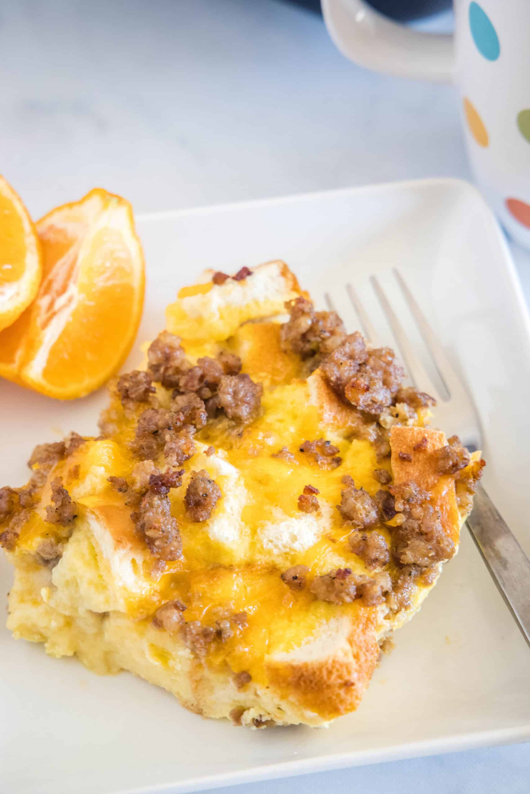 A serving of sourdough breakfast casserole next to orange slices and a fork on a white square plate.