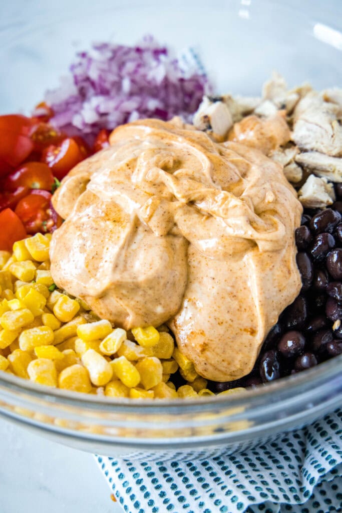 Close up of mayo and taco seasoning dressing in a mixing bowl on top of corn, tomatoes, onions, chicken, and beans