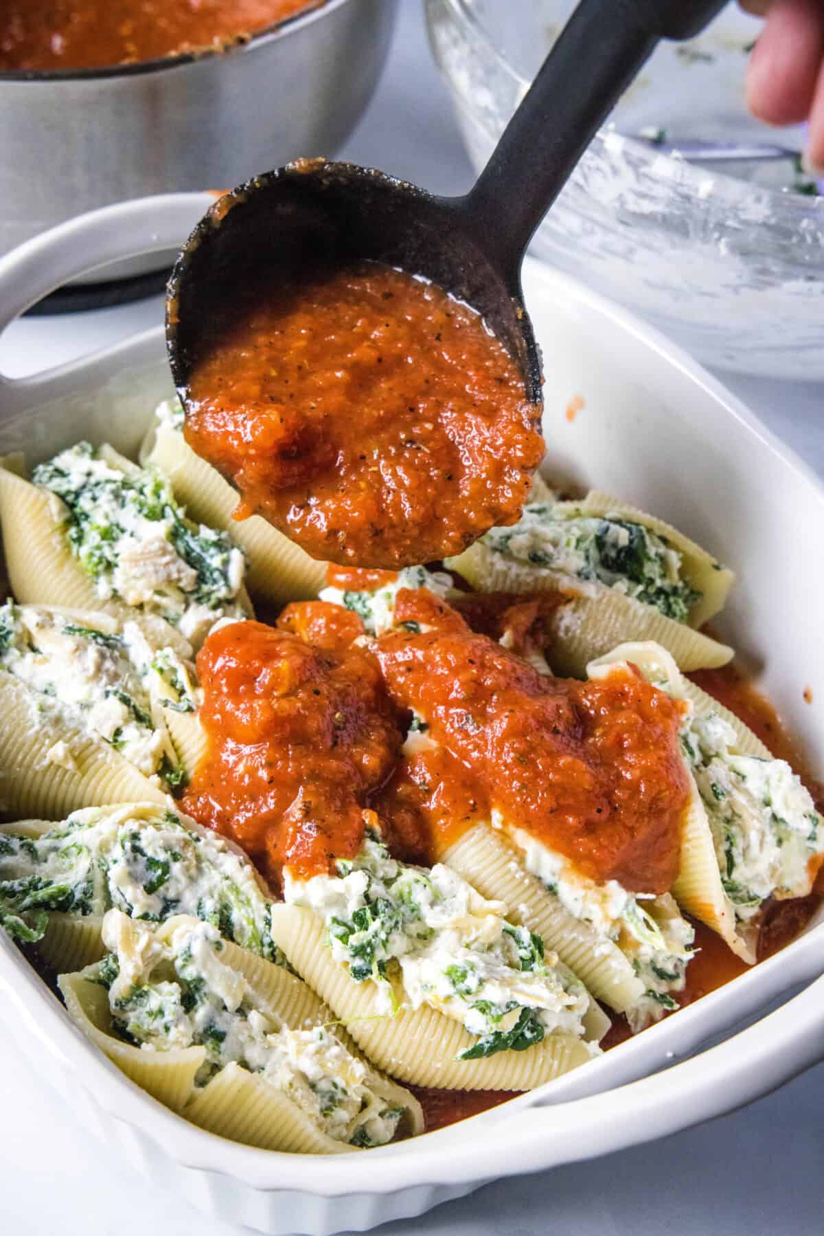 spooning red sauce over stuffed shells in a casserole dish