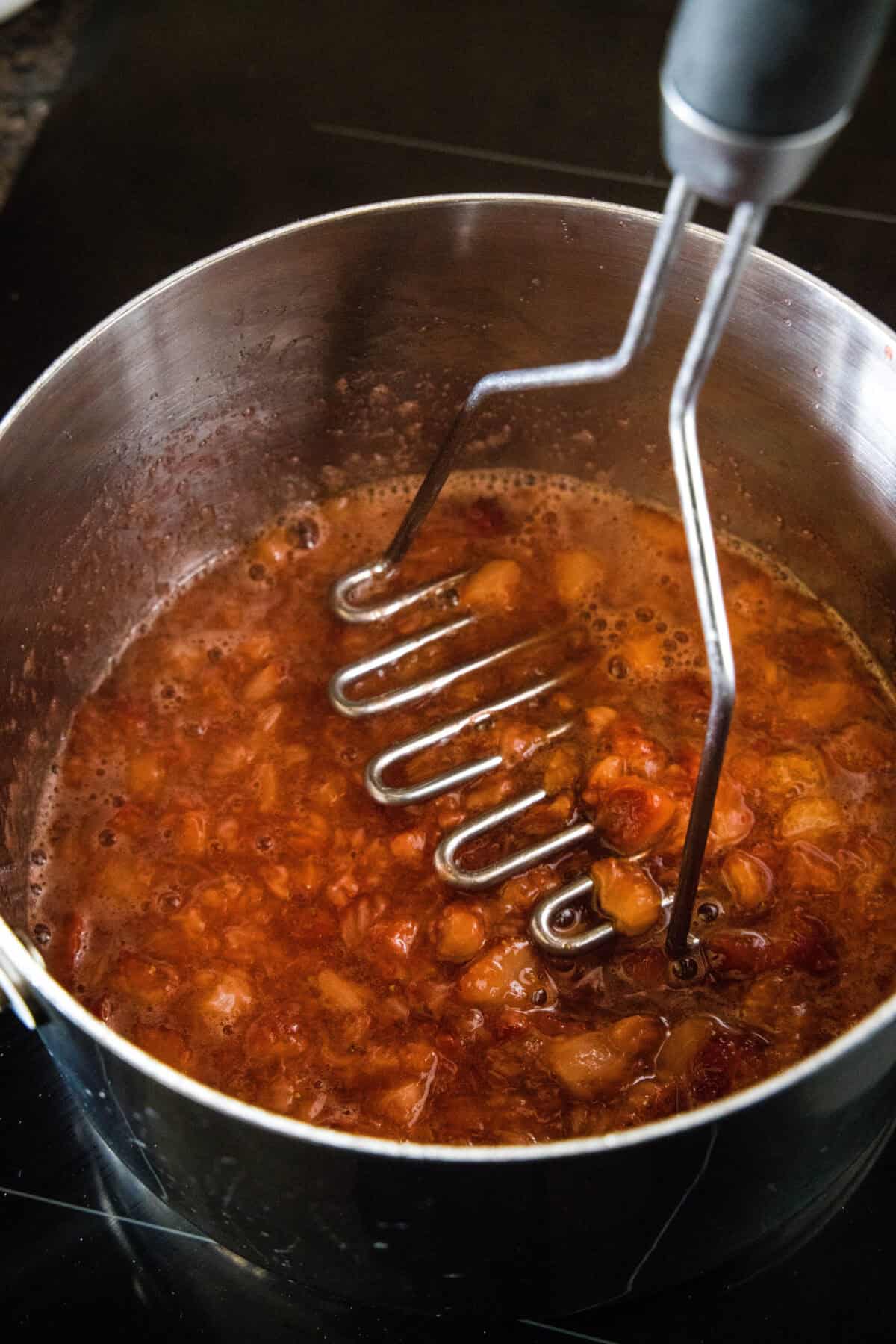 A potato masher being used to mash simmered strawberries in a saucepan.