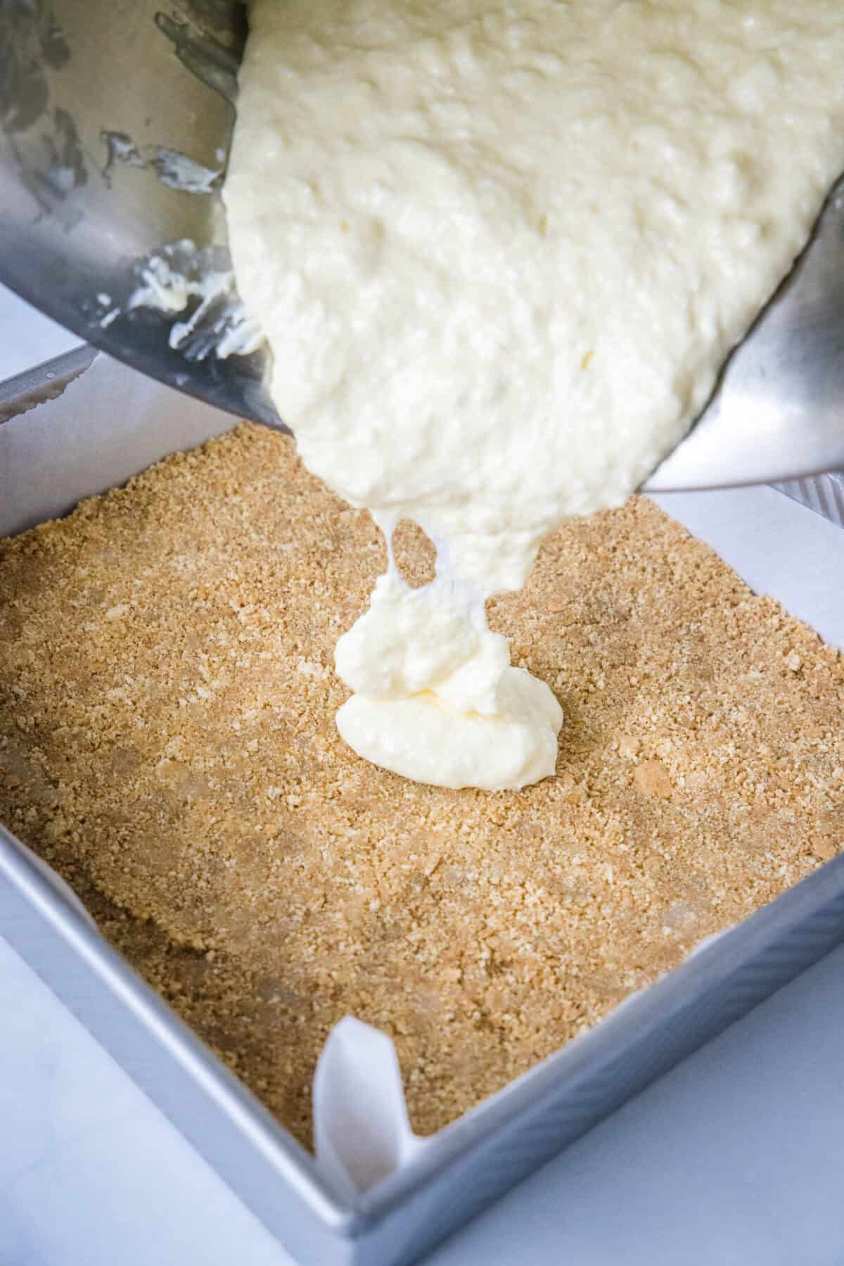 Cheesecake filling being poured over a prepared graham cracker crust in a baking dish.