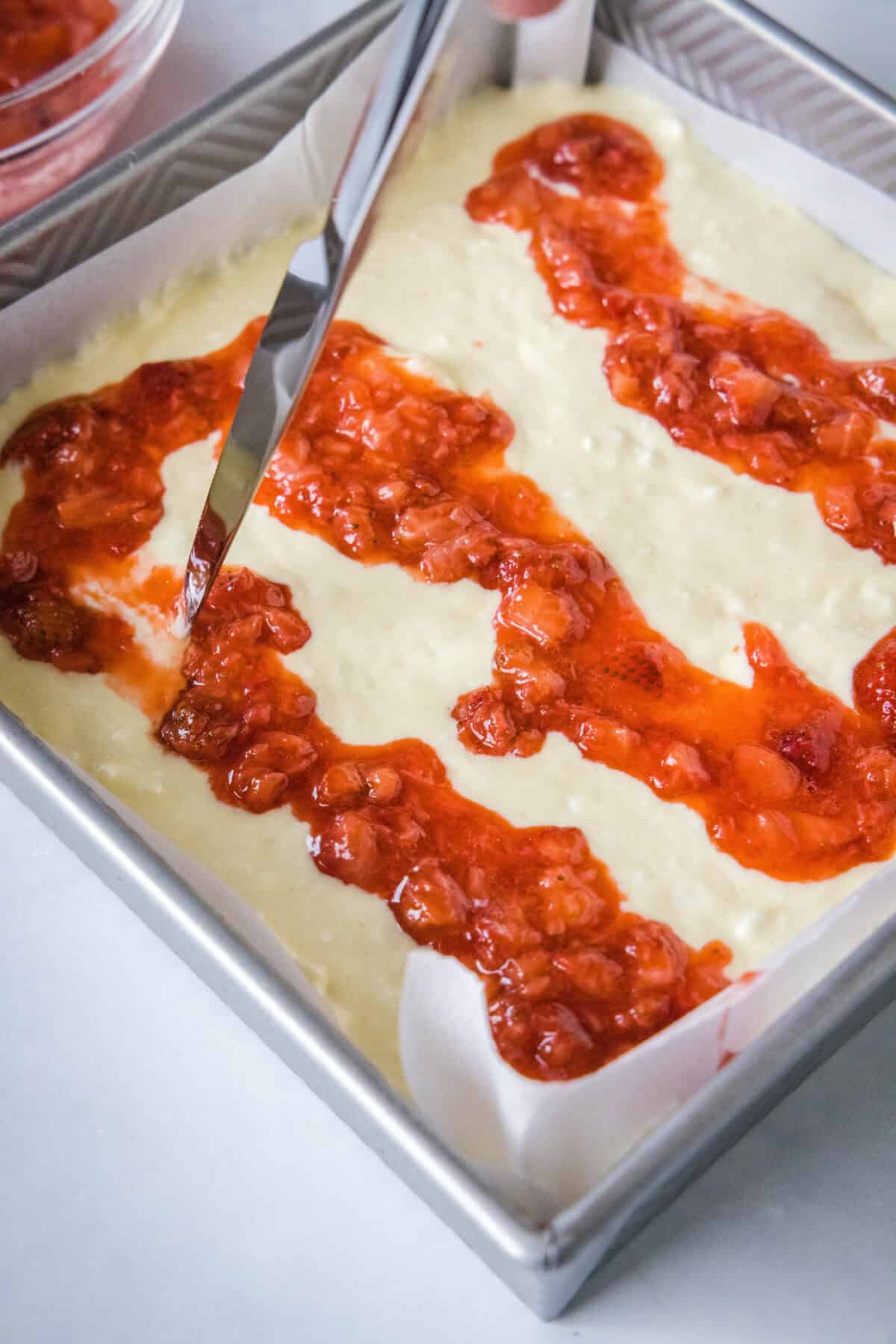 A knife being used to swirl strawberry compote into cheesecake filling.