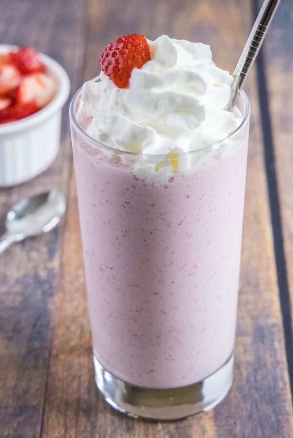 Wide angle view of a strawberry milkshake topped with whipped cream, next to a spoon and a bowl of strawberries.