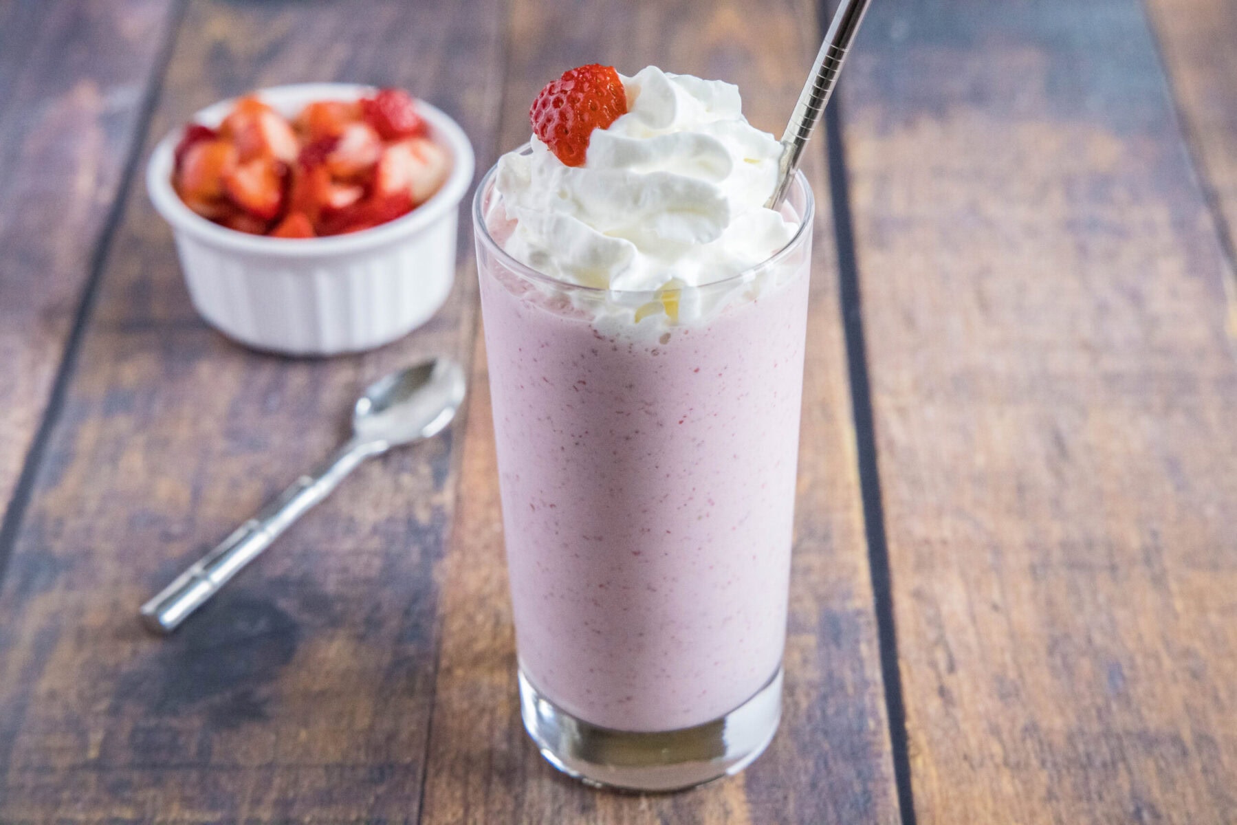 Wide angle view of a strawberry milkshake topped with whipped cream, next to a spoon and a bowl of strawberries.