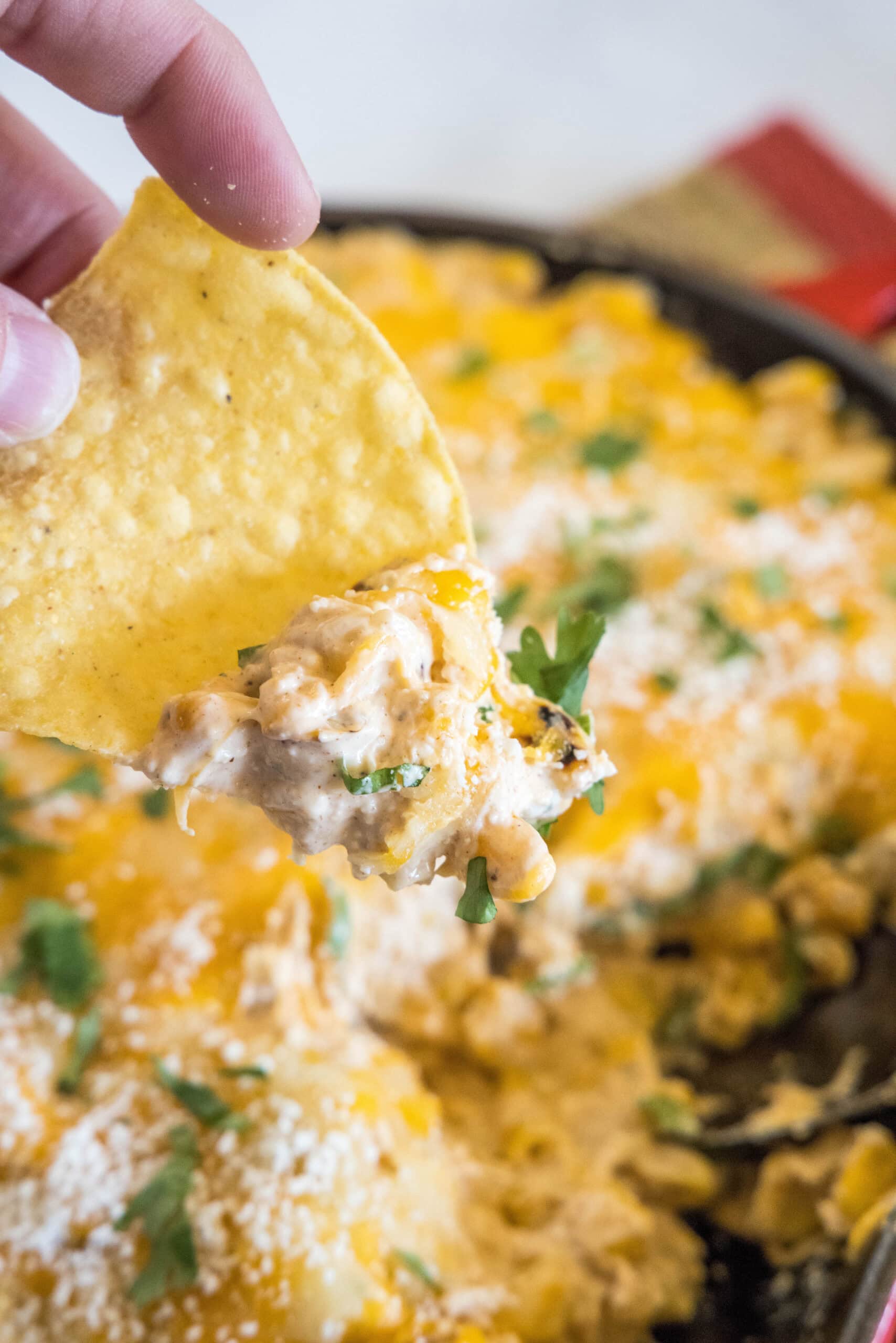 A hand dipping a tortilla chip into a skillet of cheesy street corn dip.
