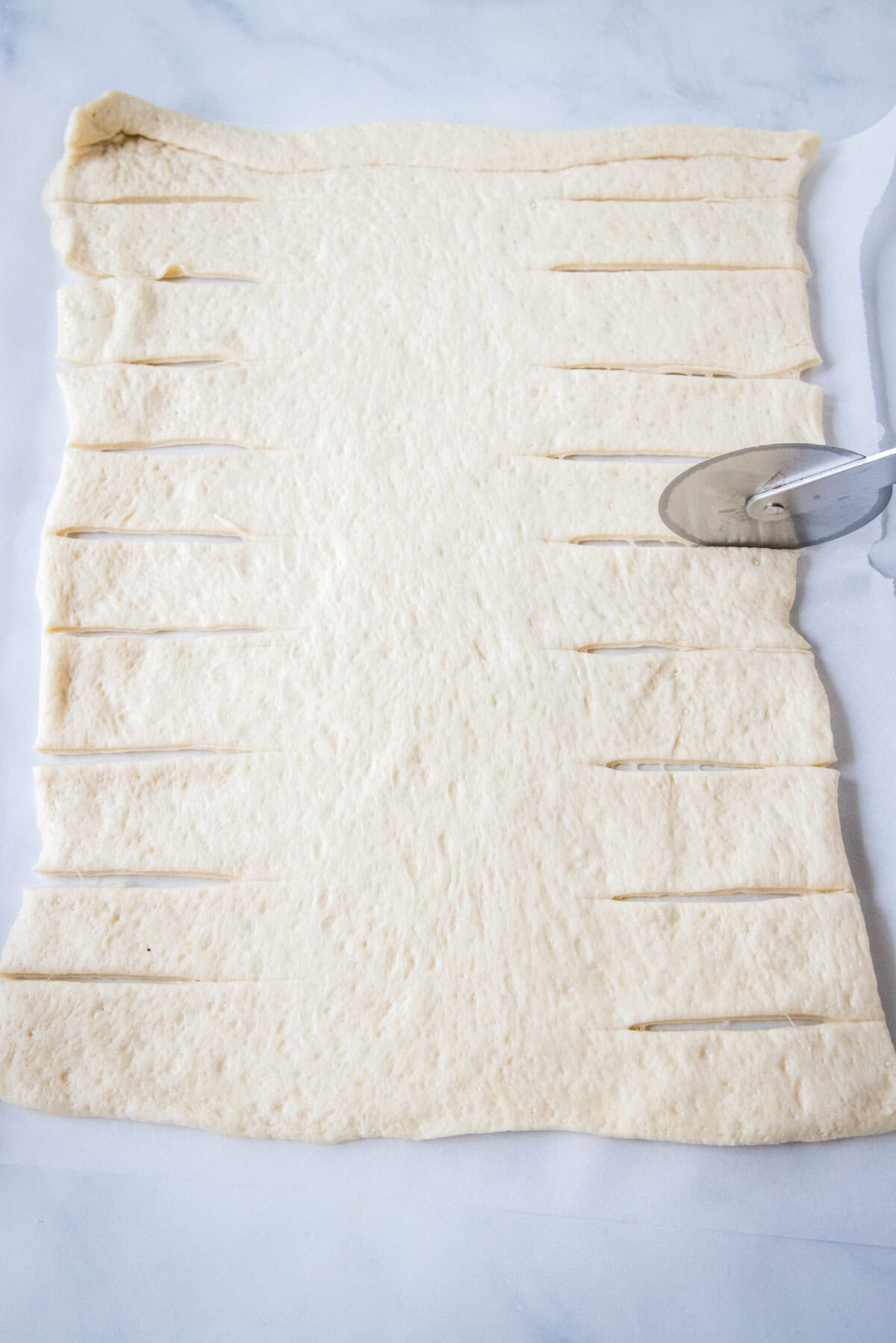 A pizza cutter is used to cut the long sides of a dough rectangle into strips.