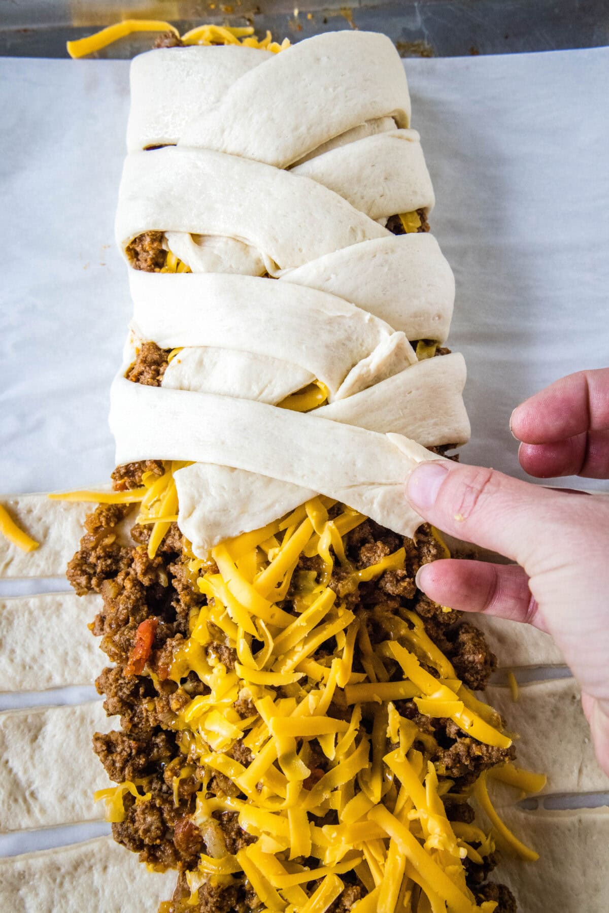 A hand braiding the dough strips over ground taco meat and cheese.