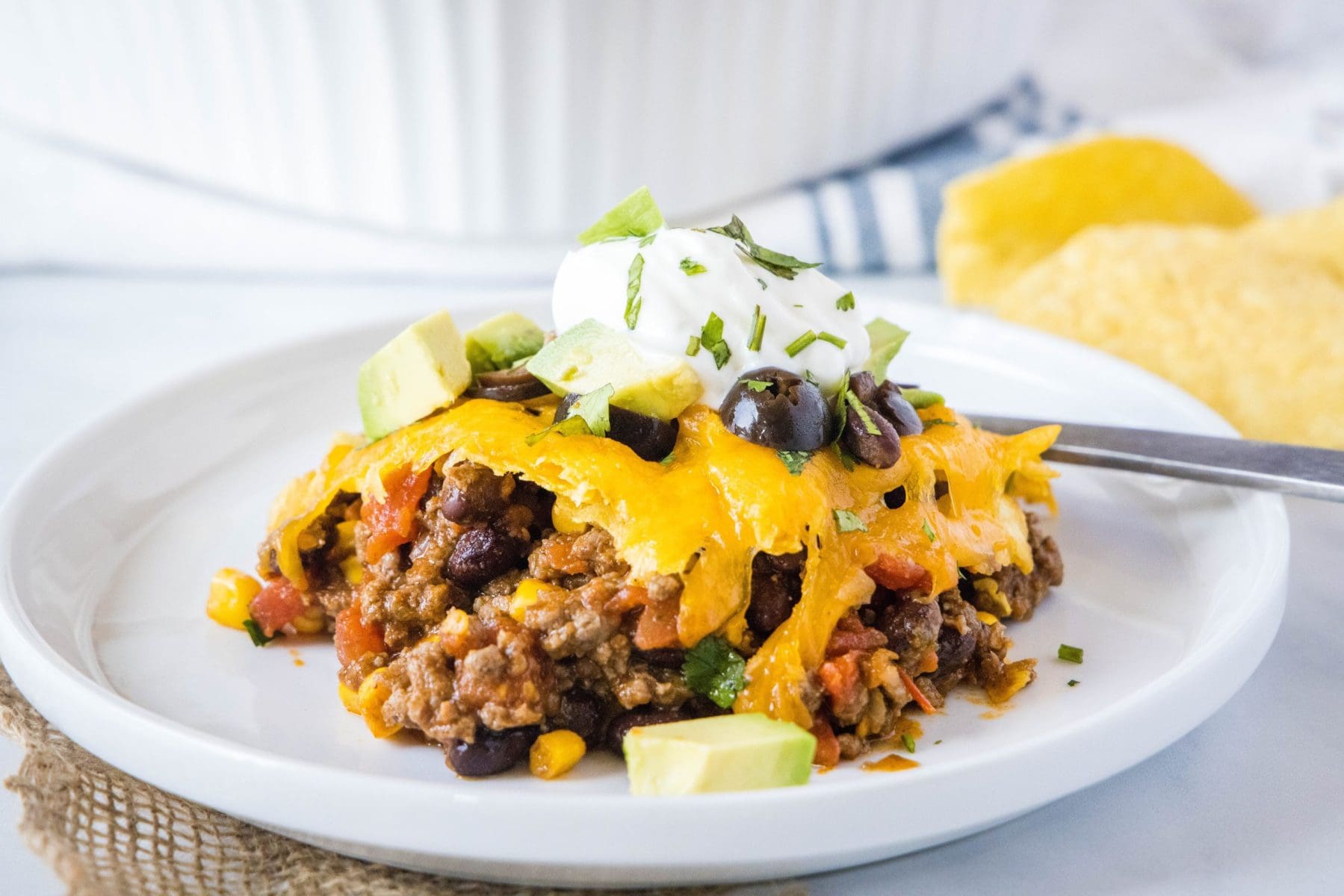 A serving of taco casserole on a plate, with a fork, topped with sour cream, cilantro, olives, and avocado, with chips in the background