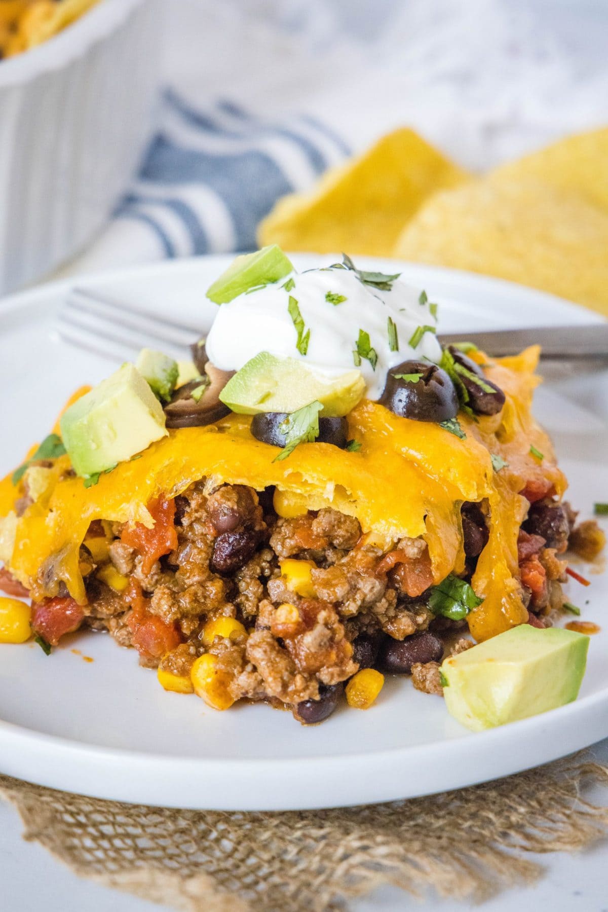 A plate with a serving of taco casserole, topped with sour cream, cilantro, olives, and avocado, with chips in the background