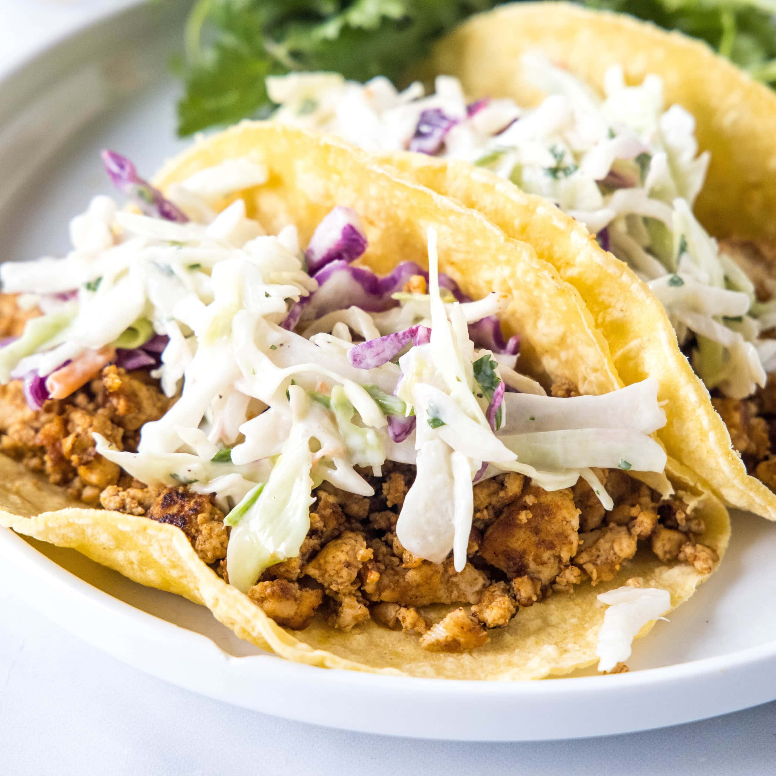 Two tofu tacos topped with Mexican coleslaw and avocado cream on a white plate.