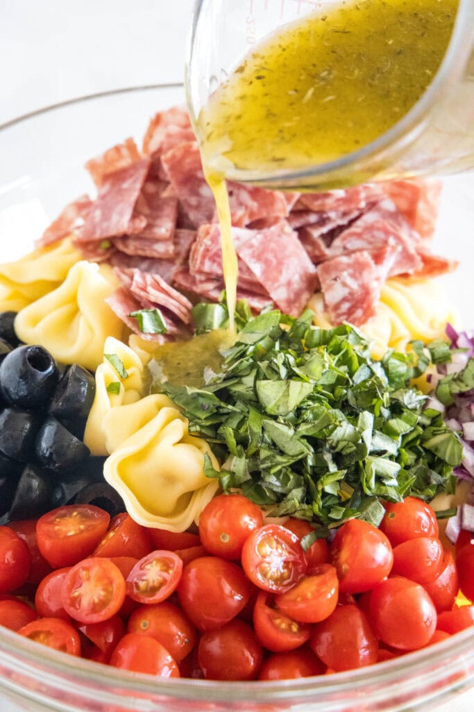 Italian dressing being poured over tortellini salad ingredients in a glass bowl.