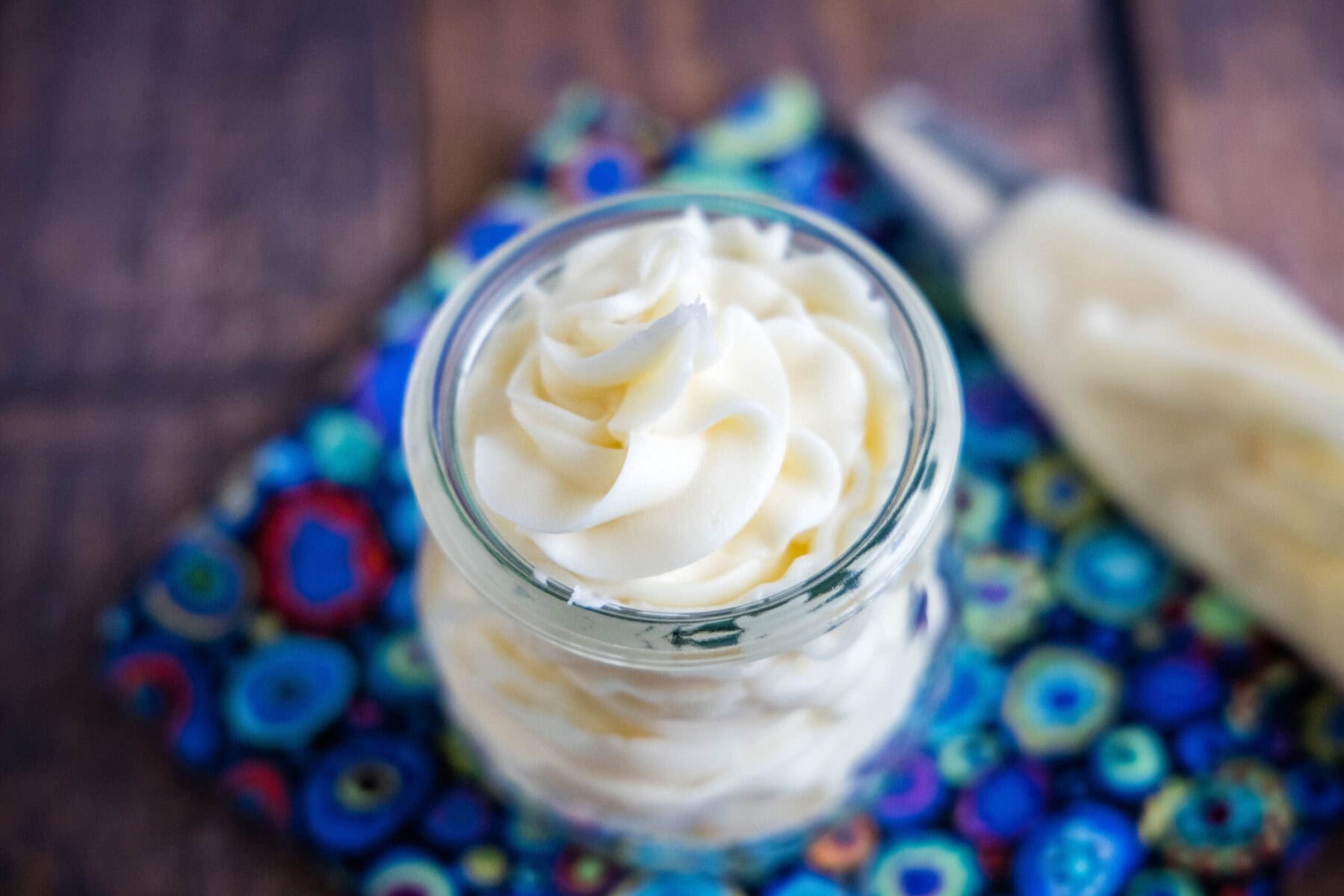 Overhead view of a jar of vanilla buttercream frosting on a kitchen towel, next to a piping bag