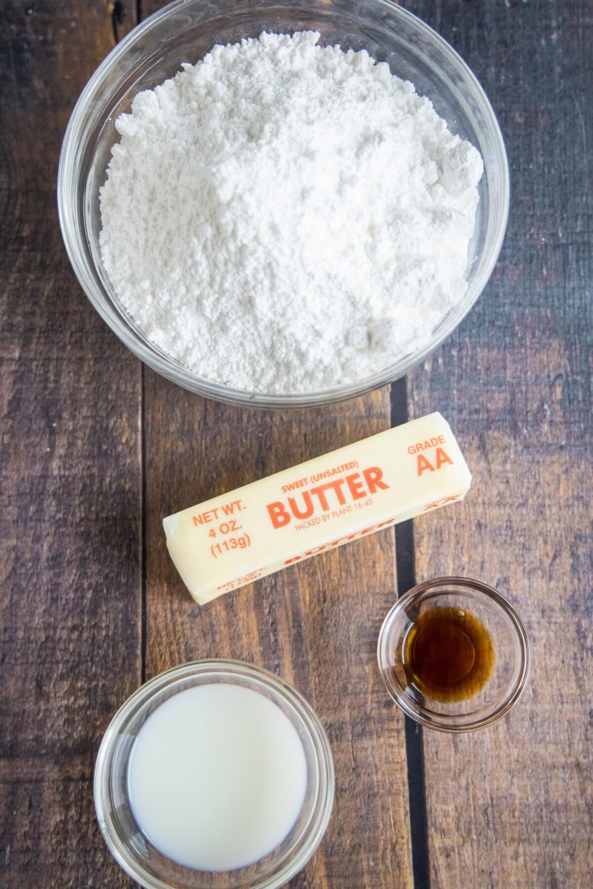 Overhead view of the ingredients needed for vanilla frosting: a bowl of powdered sugar, a bowl of milk, a bowl of vanilla, and a stick of butter