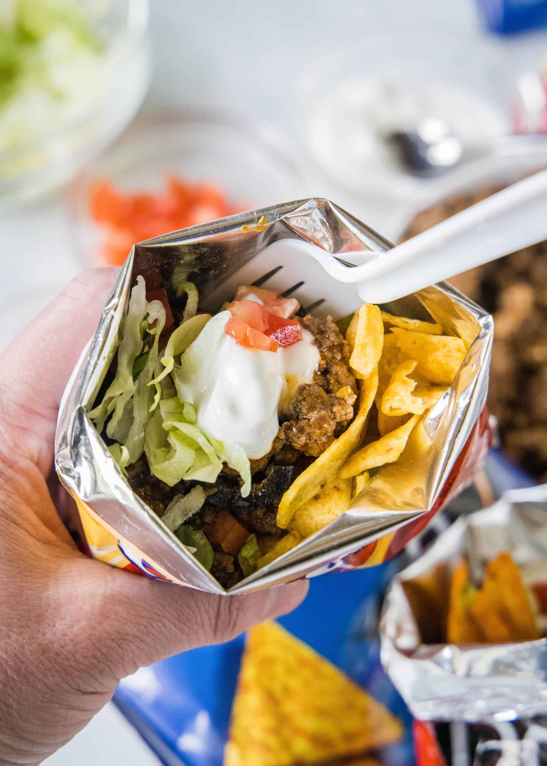 A hand holding a walking taco topped wth lettuce, tomatoes, and sour cream inside a chip bag with a fork.