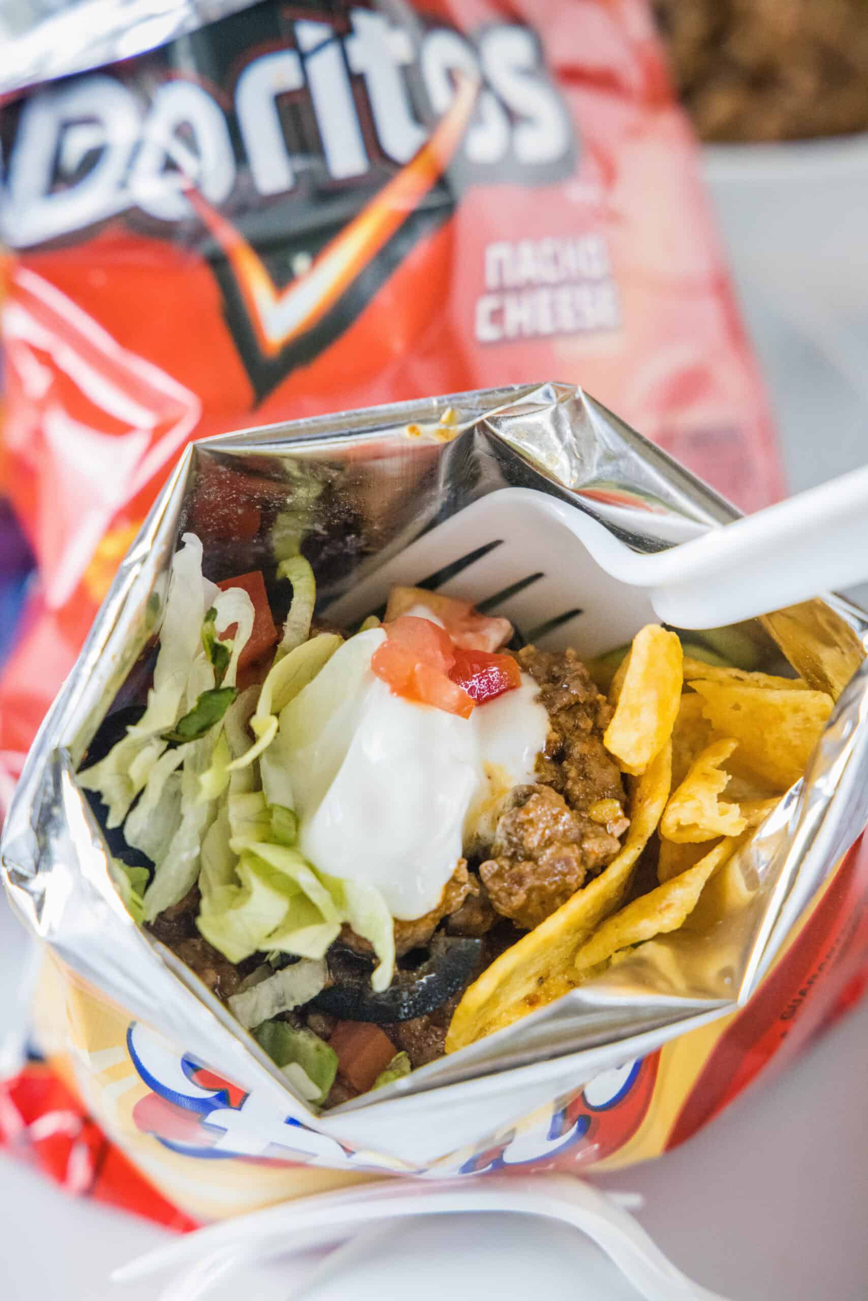 A walking taco topped wth lettuce, tomatoes, and sour cream inside a chip bag with a fork.