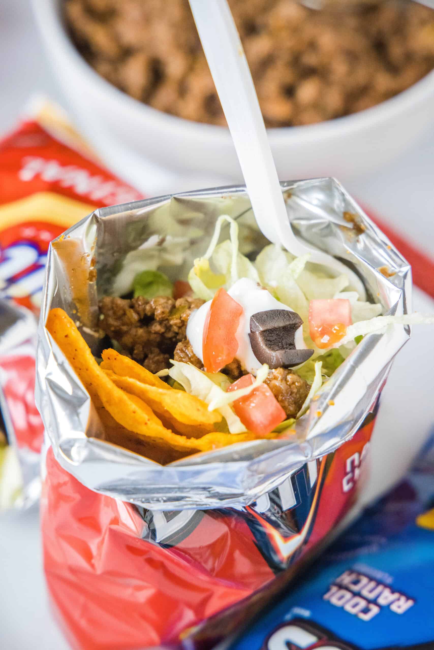 A walking taco topped wth lettuce, tomatoes, and sour cream inside a chip bag with a fork.