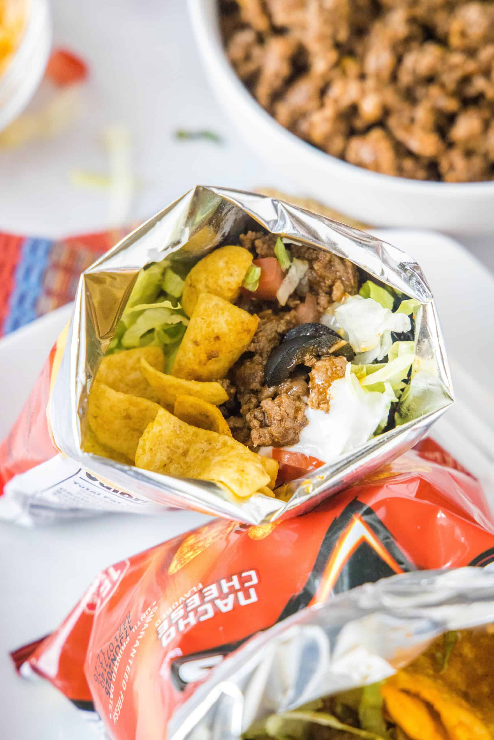 A walking taco topped wth lettuce, tomatoes, and sour cream inside a chip bag, with a bowl of taco meat in the background.