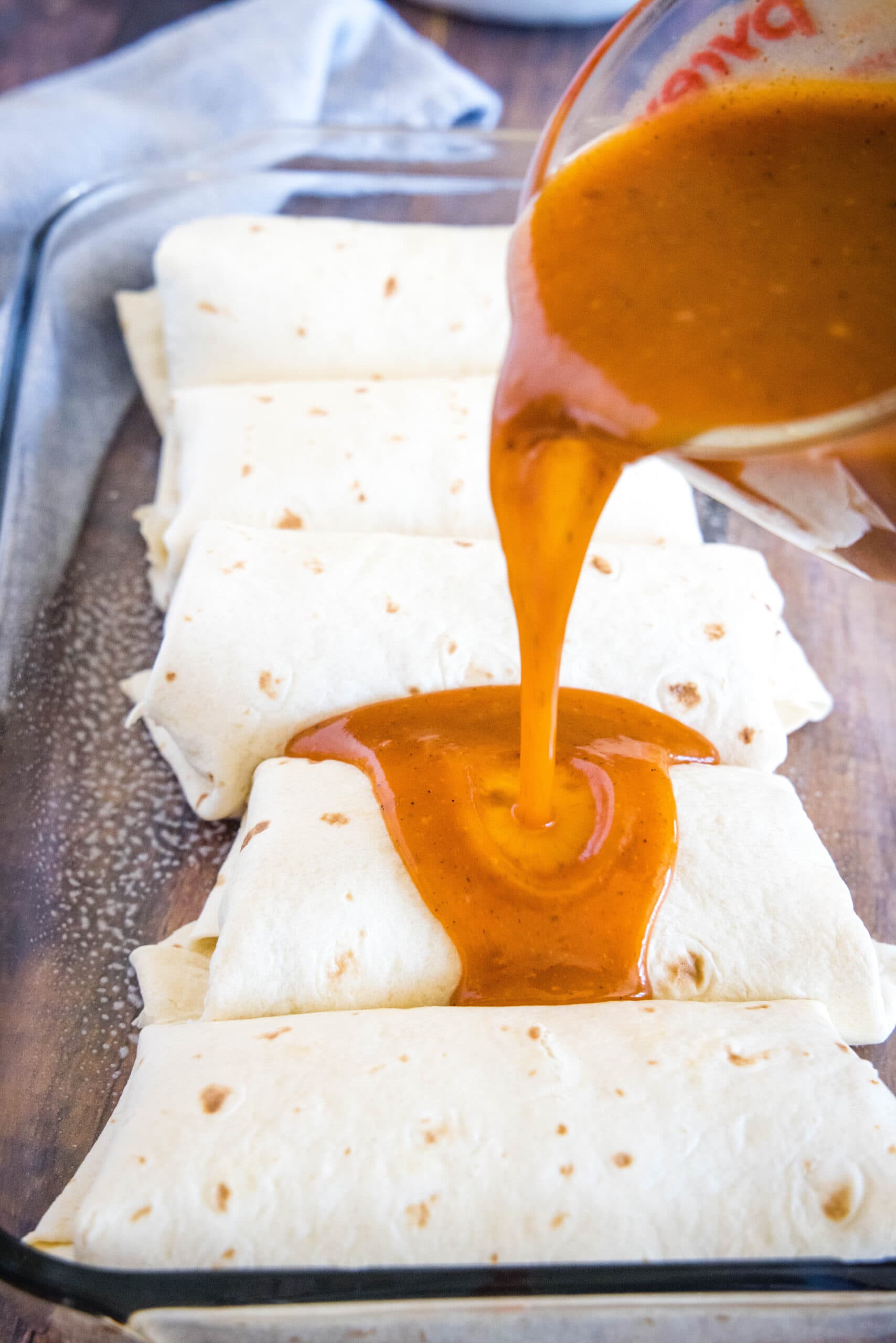 Enchilada sauce being poured over burritos in a baking dish.
